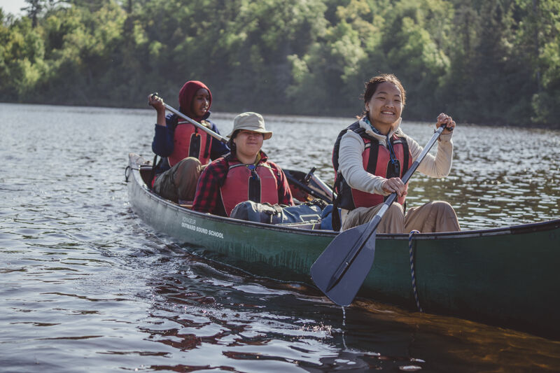 The image shows three people in a canoe on a lake. They are all wearing life jackets and holding paddles, suggesting they are paddling. The person in the front is smiling and looking towards the camera. The background features trees, indicating a natural setting. The water is calm, and the overall scene suggests a recreational activity like canoeing or kayaking.